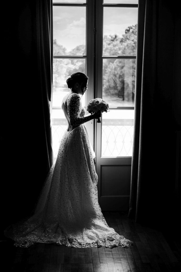 Une mariée pensive avec un bouquet de fleurs devant une fenêtre au château de Quincey en Bourgogne