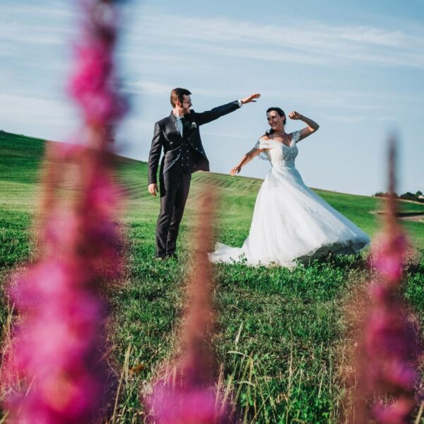 Dans le Doubs, un couple de mariés qui dansent dans un champs, avec des fleurs roses au premier plan.